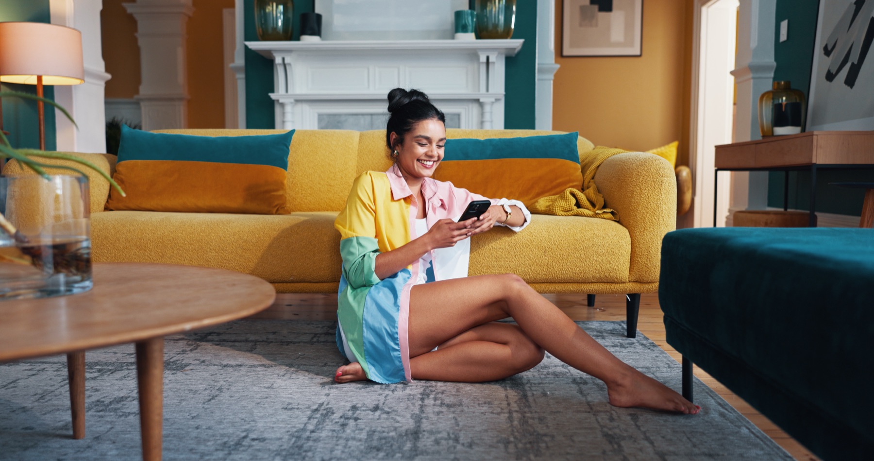 Une femme souriante consulte son téléphone dans son salon, devant un canapé jaune.